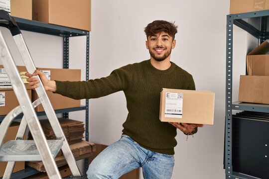 Young arab man ecommerce business worker holding package standing on ladder at office