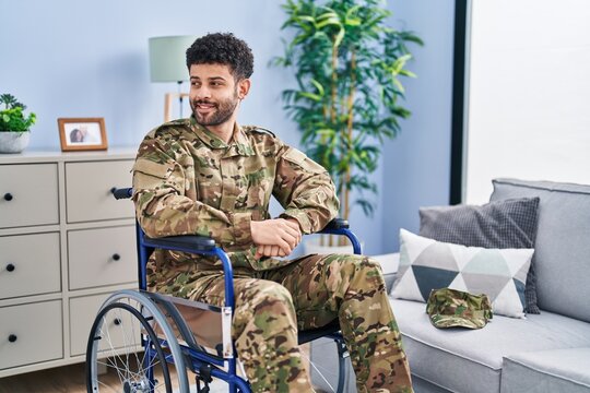 Arab man wearing camouflage army uniform sitting on wheelchair looking away to side with smile on face, natural expression. laughing confident.