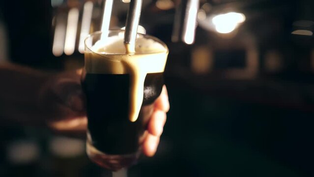 the bartender pours beer into a glass. Close-up view of barman's hand filling glass with dark beer, at bar table, at pub. Pouring beer into mug, at beer bar
