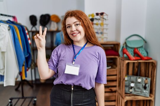 Young redhead woman working as manager at retail boutique showing and pointing up with fingers number two while smiling confident and happy.