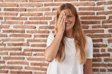 Young caucasian woman standing over bricks wall covering one eye with hand, confident smile on face and surprise emotion.