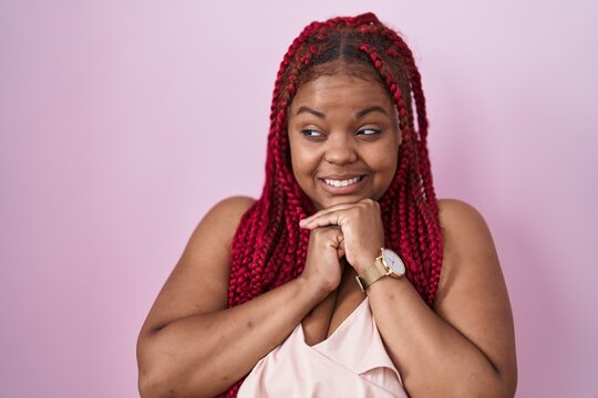 African american woman with braided hair standing over pink background laughing nervous and excited with hands on chin looking to the side