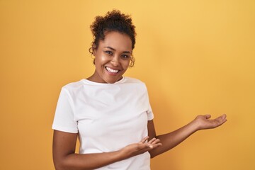 Young hispanic woman with curly hair standing over yellow background inviting to enter smiling natural with open hand