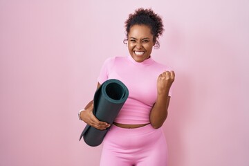 Young hispanic woman with curly hair holding yoga mat over pink background very happy and excited doing winner gesture with arms raised, smiling and screaming for success. celebration concept.