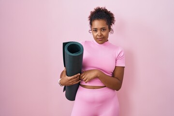 Young hispanic woman with curly hair holding yoga mat over pink background looking sleepy and...