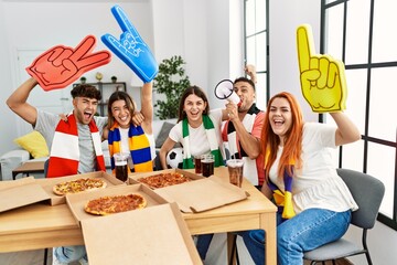 Group of young friends watching and supporting soccer match eating pizza at home.