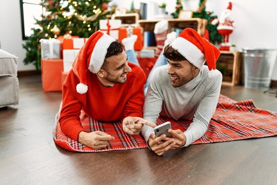 Two hispanic men couple using smartphone lying by christmas tree at home