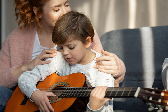 Happy mother teaching her son how to play the guitar in their living room on sunny day