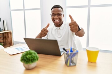 Young african man working at the office using computer laptop approving doing positive gesture with hand, thumbs up smiling and happy for success. winner gesture.