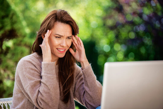 Middle aged woman sitting outdoor with laptop on desk and touching her temples