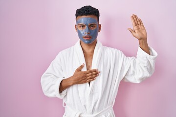 Young hispanic man wearing beauty face mask and bath robe swearing with hand on chest and open palm, making a loyalty promise oath