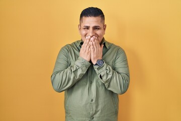 Hispanic young man standing over yellow background laughing and embarrassed giggle covering mouth with hands, gossip and scandal concept