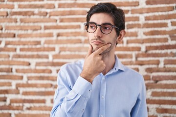 Young hispanic man standing over brick wall background with hand on chin thinking about question, pensive expression. smiling with thoughtful face. doubt concept.