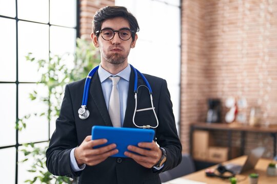Young hispanic man wearing doctor stethoscope at the office puffing cheeks with funny face. mouth inflated with air, catching air.