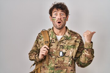 Hispanic young man wearing camouflage army uniform surprised pointing with hand finger to the side, open mouth amazed expression.