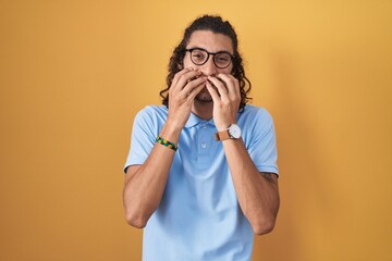 Young hispanic man standing over yellow background laughing and embarrassed giggle covering mouth...