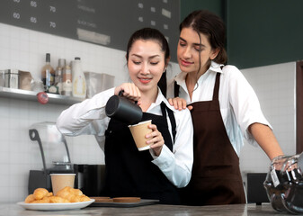 Young barista and waitress women standing at cafeteria counter bar preparing cappuccino in take away coffee cup for delivery. Two lesbian women couple manage cafe business together. LGBTQ lifestyle.