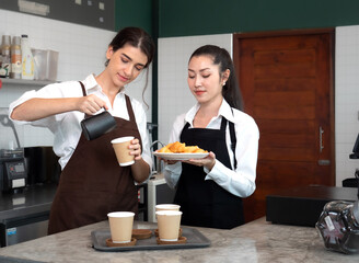 Young barista and waitress women standing at cafeteria counter bar preparing cappuccino in take away coffee cup for delivery. Two lesbian women couple manage cafe business together. LGBTQ lifestyle.