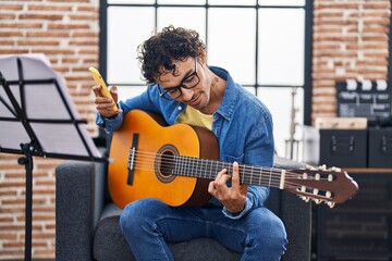 Young hispanic man musician playing classical guitar using smartphone at music studio