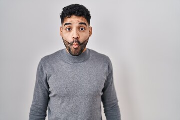 Hispanic man with beard standing over white background making fish face with lips, crazy and comical gesture. funny expression.