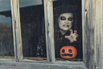 Little girl in Halloween costume of witch with pumpkin jack-o-lantern looking through window