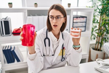 Young caucasian dentist woman holding denture and mouthwash in shock face, looking skeptical and...