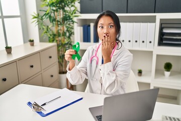 Young asian doctor woman holding support green ribbon covering mouth with hand, shocked and afraid...
