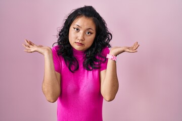 Young asian woman standing over pink background clueless and confused expression with arms and hands raised. doubt concept.