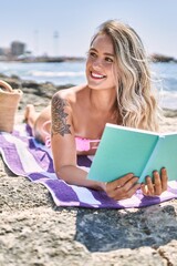 Young blonde girl reading book lying on the towel at the beach.