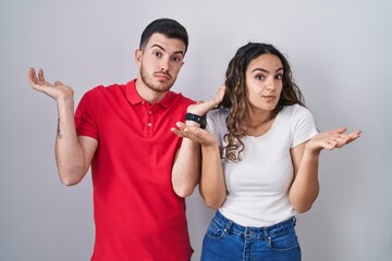 Young hispanic couple standing over isolated background clueless and confused expression with arms and hands raised. doubt concept.