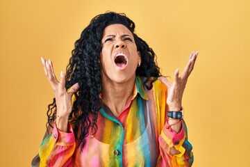 Middle age hispanic woman standing over yellow background crazy and mad shouting and yelling with aggressive expression and arms raised. frustration concept.