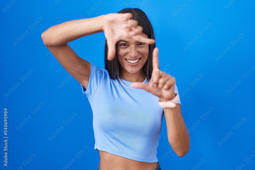 Poster brunette young woman standing over blue background smiling making frame with hands and fingers with 