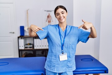 Obraz premium Young hispanic woman wearing physiotherapist uniform standing at clinic looking confident with smile on face, pointing oneself with fingers proud and happy.