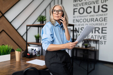 Fifty-year-old businesswoman with documents in her hands speaks on a mobile phone in a modern...