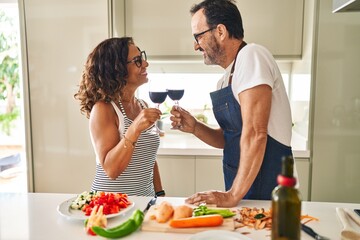 Middle age hispanic couple cooking and toasting with wine at kitchen