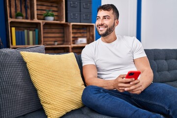 Young hispanic man using smartphone sitting on sofa at home