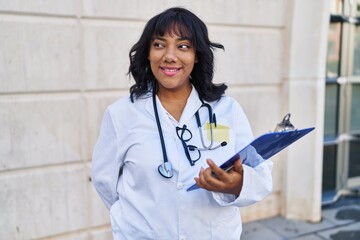 Young beautiful latin woman doctor smiling confident holding clipboard at hospital