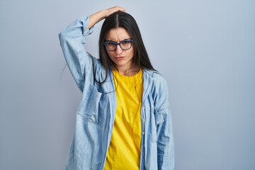 Young hispanic woman standing over blue background confuse and wondering about question. uncertain with doubt, thinking with hand on head. pensive concept.