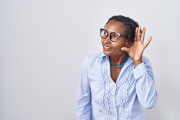 African woman with dreadlocks standing over white background wearing glasses smiling with hand over ear listening an hearing to rumor or gossip. deafness concept.