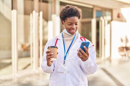 African american woman wearing doctor uniform and medical mask using smartphone drinking coffee at street