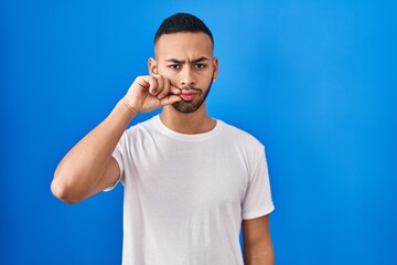Young hispanic man standing over blue background mouth and lips shut as zip with fingers. secret...