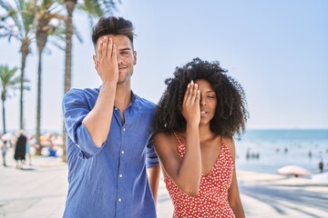 Young interracial couple outdoors on a sunny day covering one eye with hand, confident smile on face and surprise emotion.