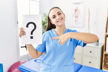 Young physiotherapist woman holding question mark smiling happy pointing with hand and finger