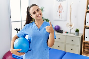 Young physiotherapist woman holding pilates ball at medical clinic smiling happy and positive, thumb up doing excellent and approval sign