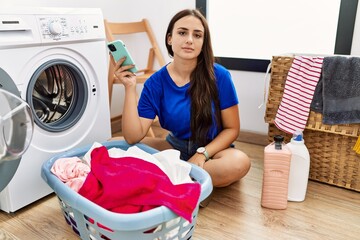 Young brunette woman doing laundry using smartphone relaxed with serious expression on face. simple and natural looking at the camera.