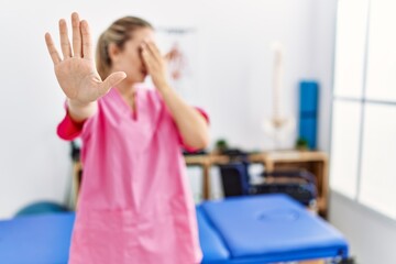 Young blonde woman working at pain recovery clinic covering eyes with hands and doing stop gesture with sad and fear expression. embarrassed and negative concept.