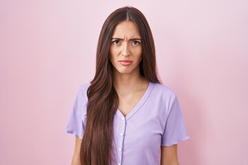 Young hispanic woman with long hair standing over pink background skeptic and nervous, frowning...