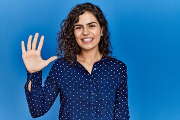 Young brunette woman with curly hair wearing casual clothes over blue background showing and pointing up with fingers number five while smiling confident and happy.