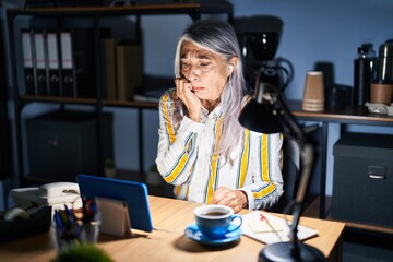 Middle age woman with grey hair working at the office at night looking stressed and nervous with hands on mouth biting nails. anxiety problem.