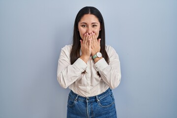 Young latin woman standing over blue background laughing and embarrassed giggle covering mouth with...